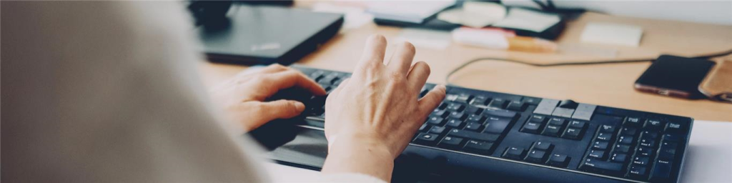 Person typing on a black computer keyboard with a wooden desk in the background, showcasing a workspace environment.