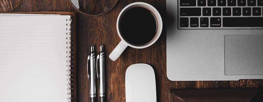 Picture of a laptop, cup of coffee, notepad and pens on a desk, viewed from above.